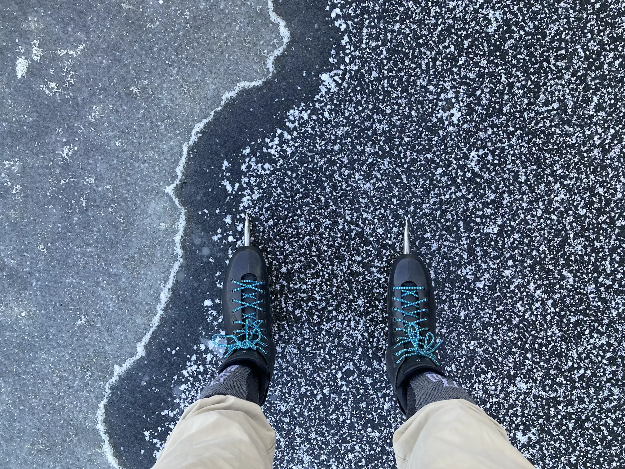 A first-person-view of a pair of legs with ice skates on an ice surface with textures of snow in various organic forms.